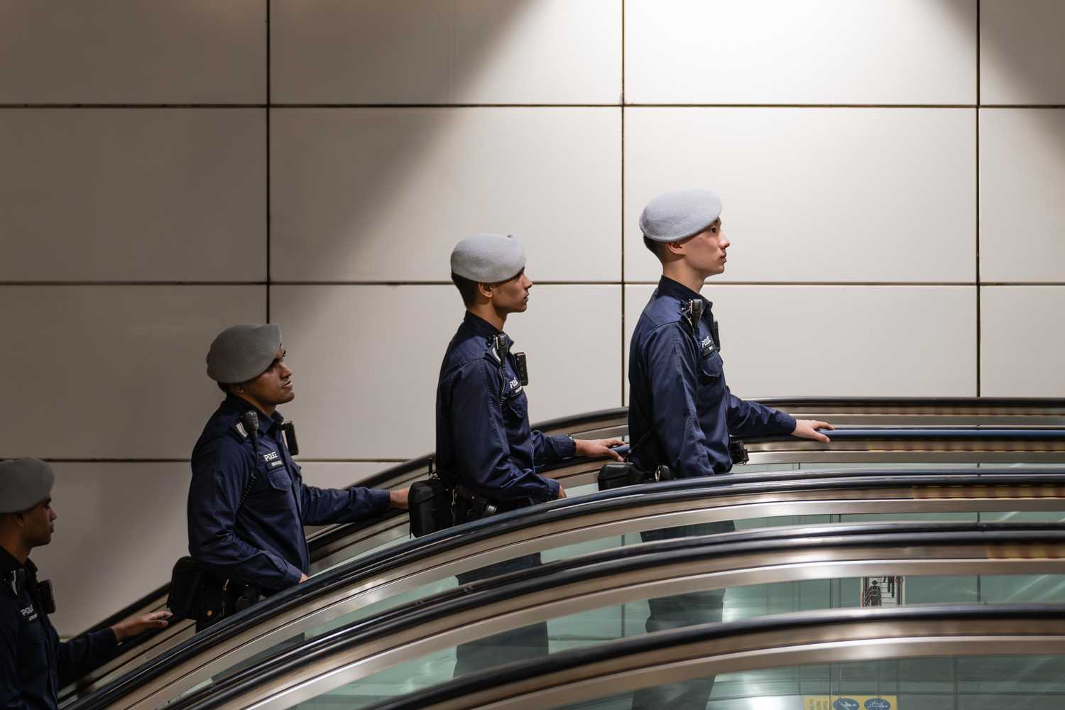 four transcom officer in blue uniform and grey beret, riding up an escalator, half appearing on the platform level in sync to the right