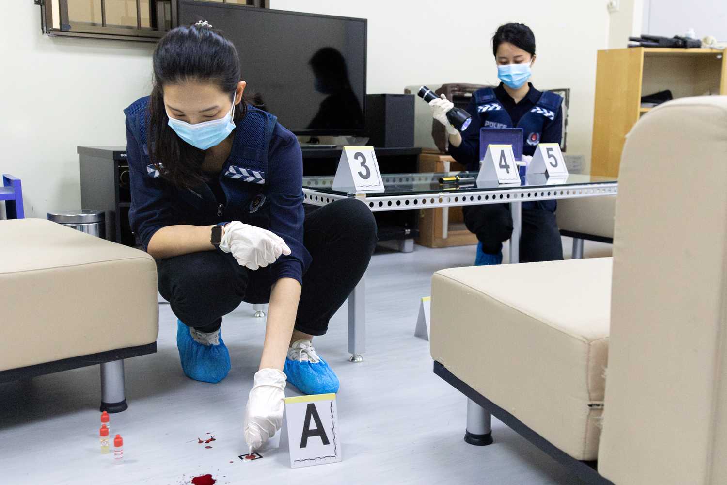 Two Forensic Specialists squatting at a mock crime scene. The first Forensic Specialist in the foreground is picking up blood stains on the floor with a cotton bud for further testing and the second Forensic Specialist in the background is shining a Forensic Light Source containing UV light on some artefacts.
