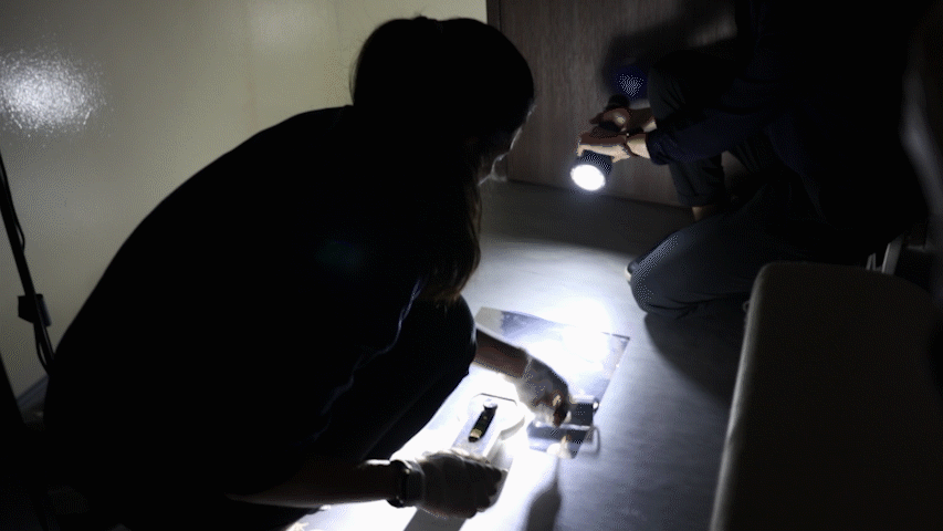 A gif showing two Forensic Specialists squatting at a mock crime scene. One is holding a flashlight so the other can see in the dark. The second Forensic Specialist is squatting on the floor and spreading aluminum foil over a black vinyl sheet as part of deploying the ESLA.