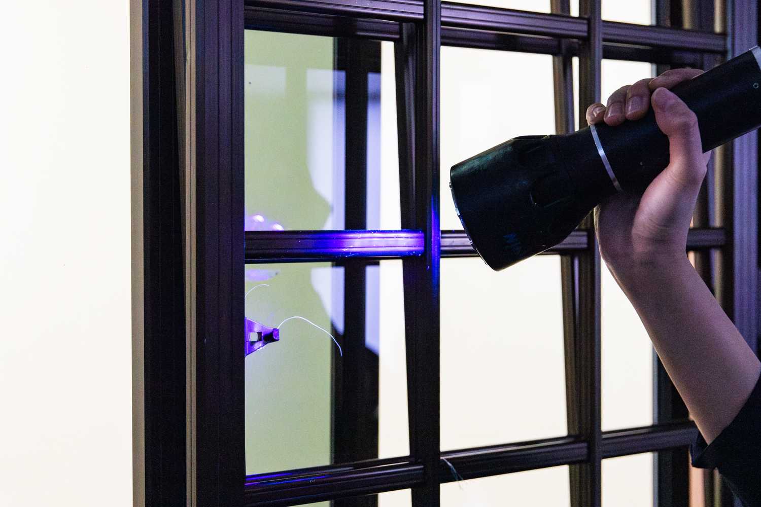 A Forensic Specialist's hand is seen holding a Forensic light source containing ultraviolet light and shining it on a window grill. Under the light of the Forensic light source, some trace evidence is seen, which is previously not visible under the naked eye and under normal light conditions.