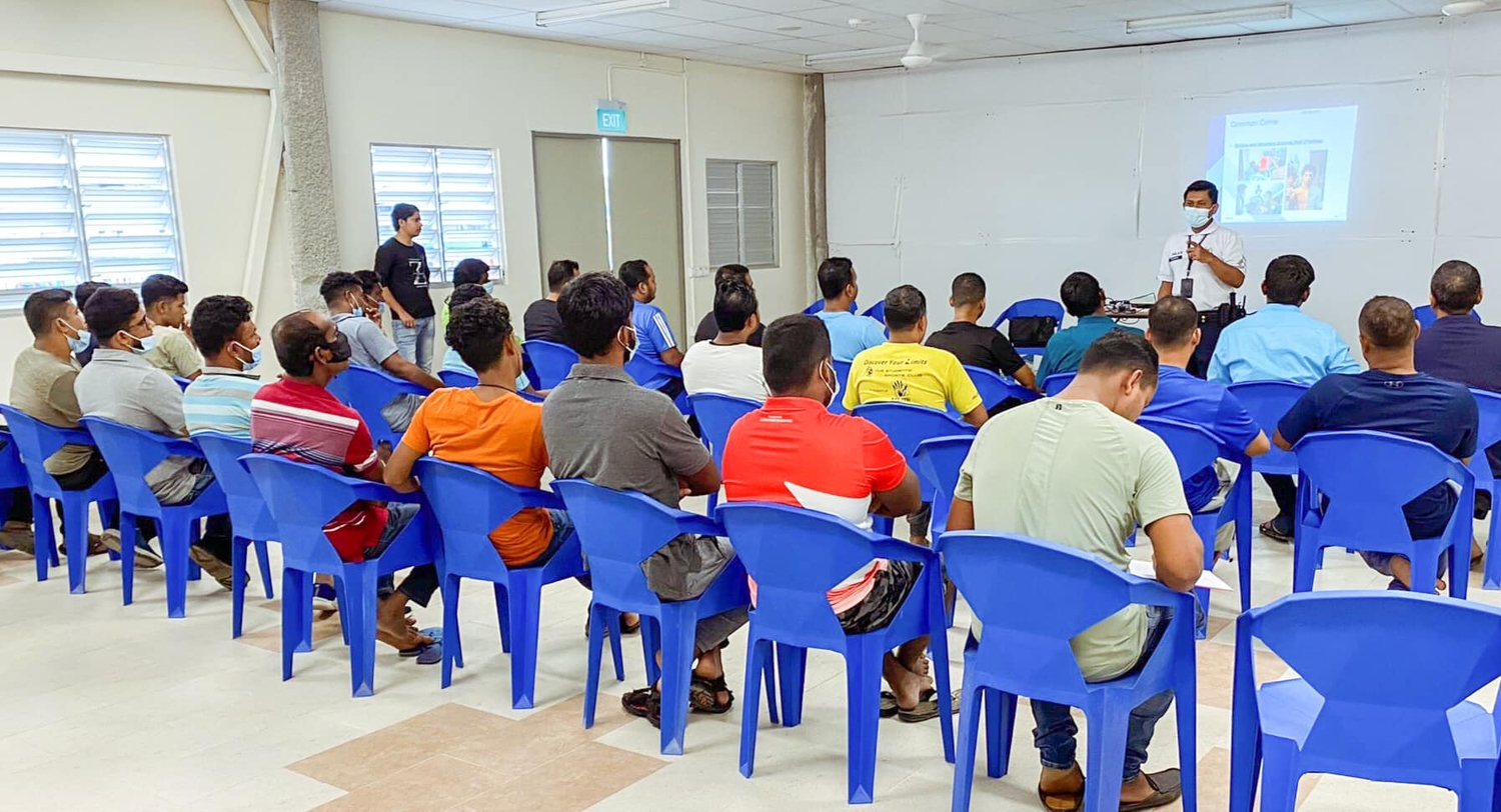 many migrant workers inside a room, sitting on blue plastic chairs and attending to a talk by a police officer on the 2pm direction of the image