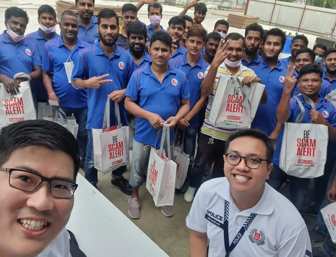 two community policing unit officers in white polo tee taking a selfie with migrant workers in the background