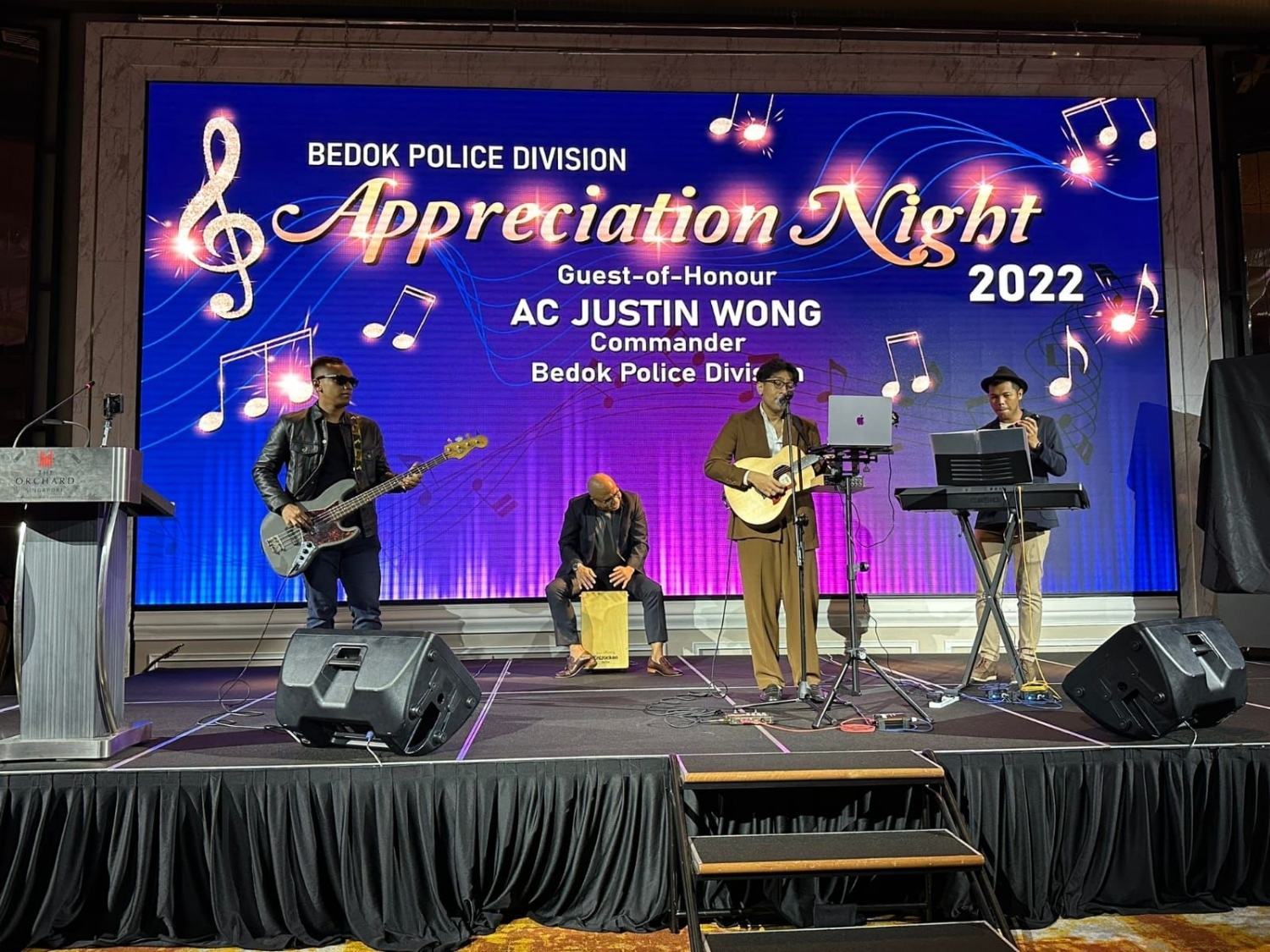 four officers on stage performing. There is a bright blue backdrop that says "Bedok Police Division Appreciation Night 2022" in the background.