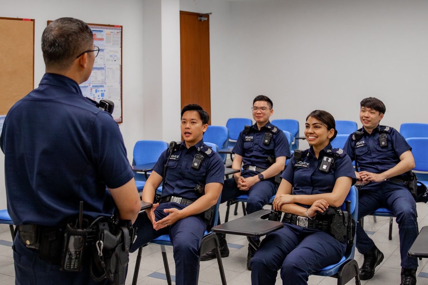 a male officer in uniform speaking in a briefing room, with many officers seated and facing him