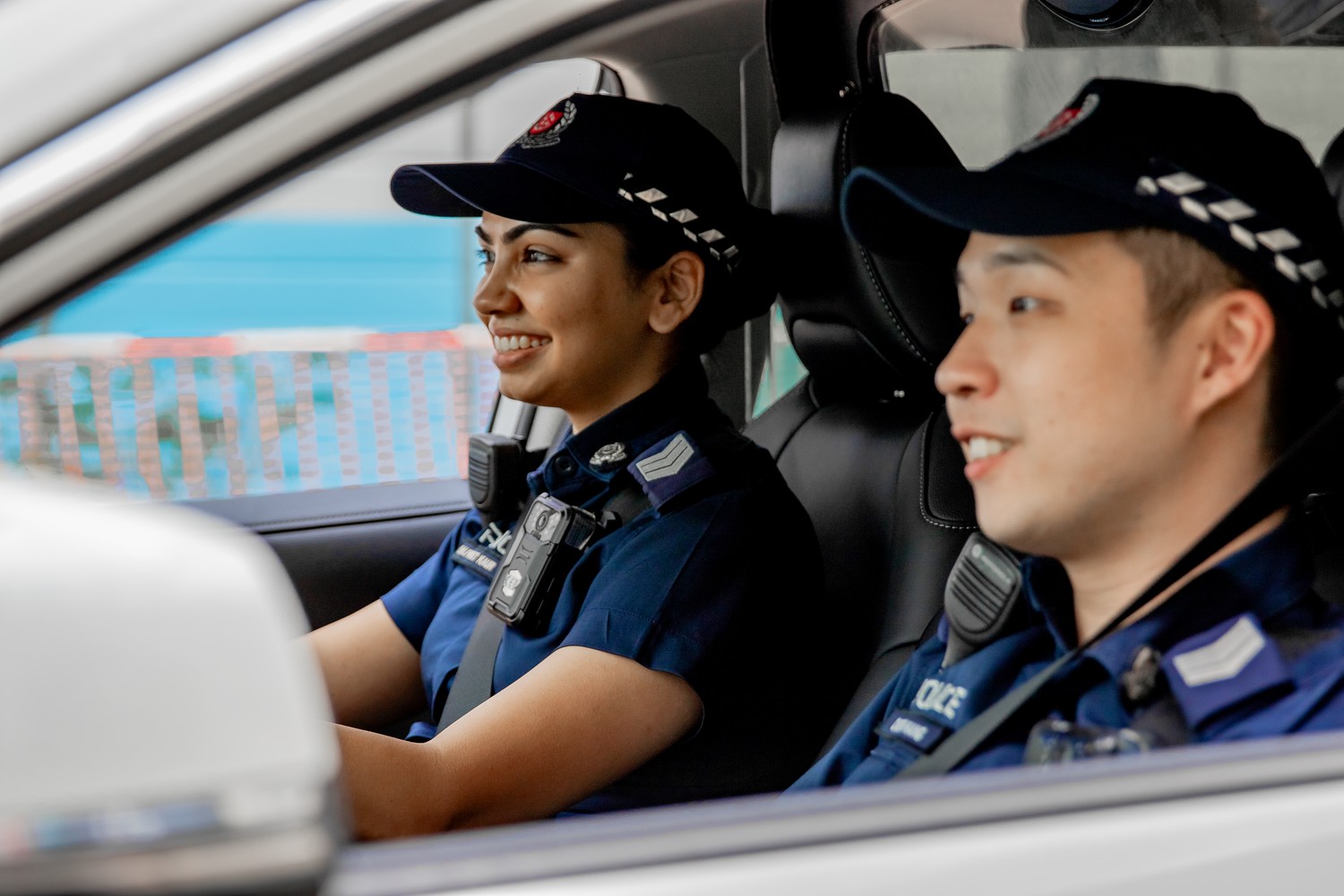 two officers in a police car looking to the left. Female officer has hands on steering wheel