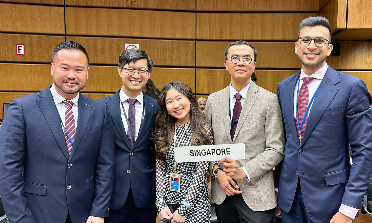 five officers in blazers and formal attire standing together, with one holding a sign that reads "Singapore"