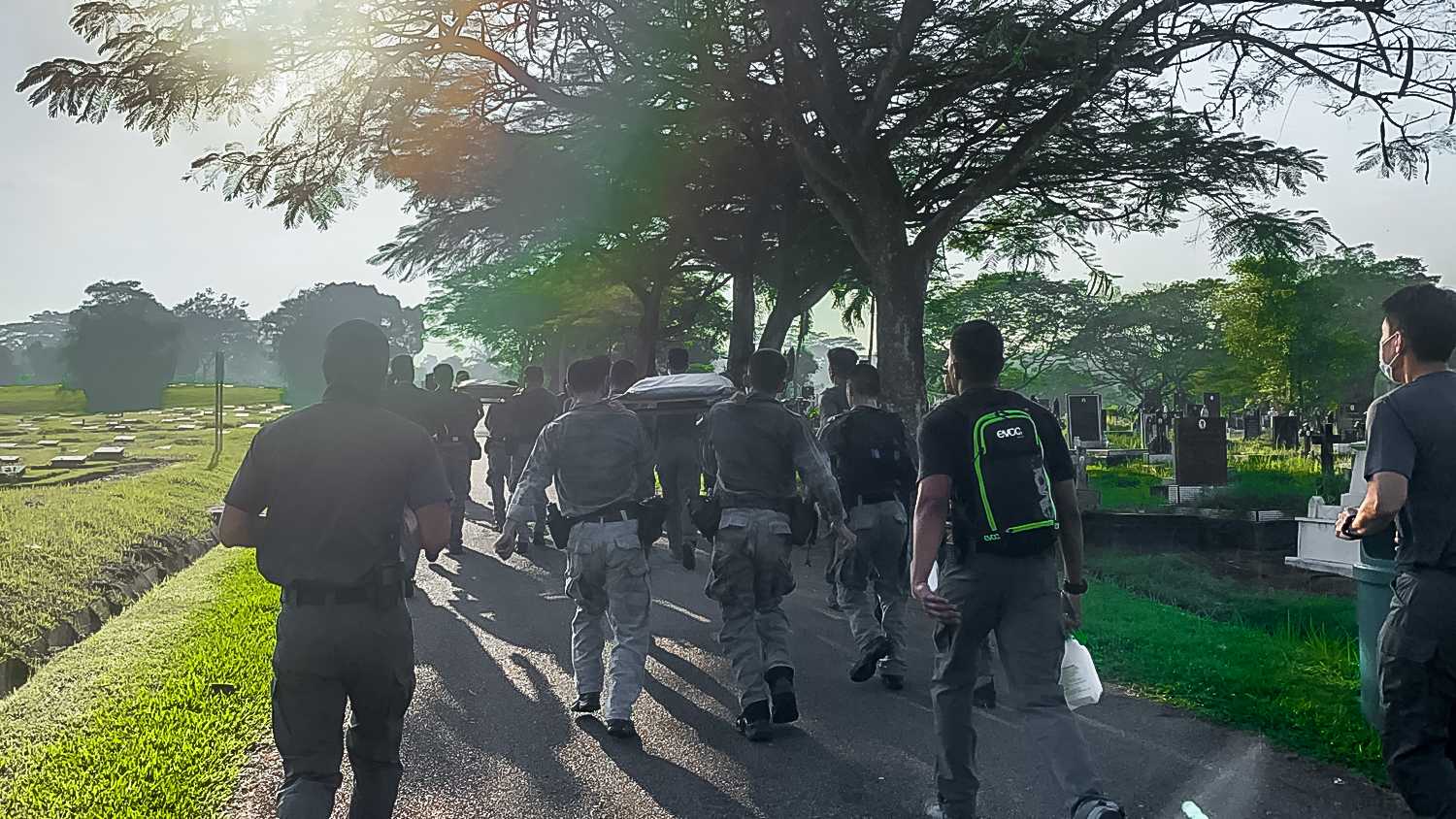 A group of people walking down a road carrying heavy load and in half uniform like they are at training.