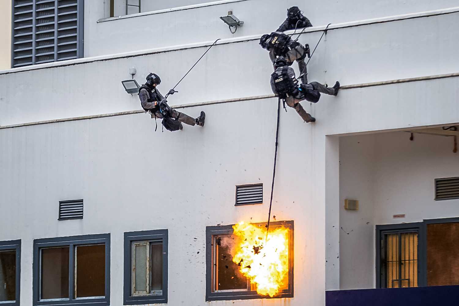 three police officers in black uniforms rappelling down a building with white walls and detonating an explosive at the window below to gain access.