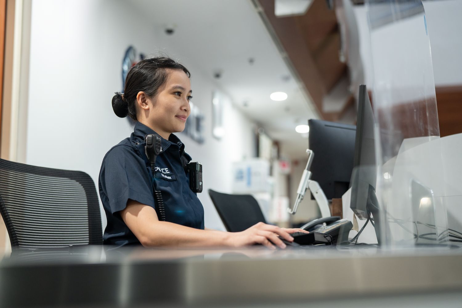 female police officer, with bunned up hair, typing at the computer located at the police front counter