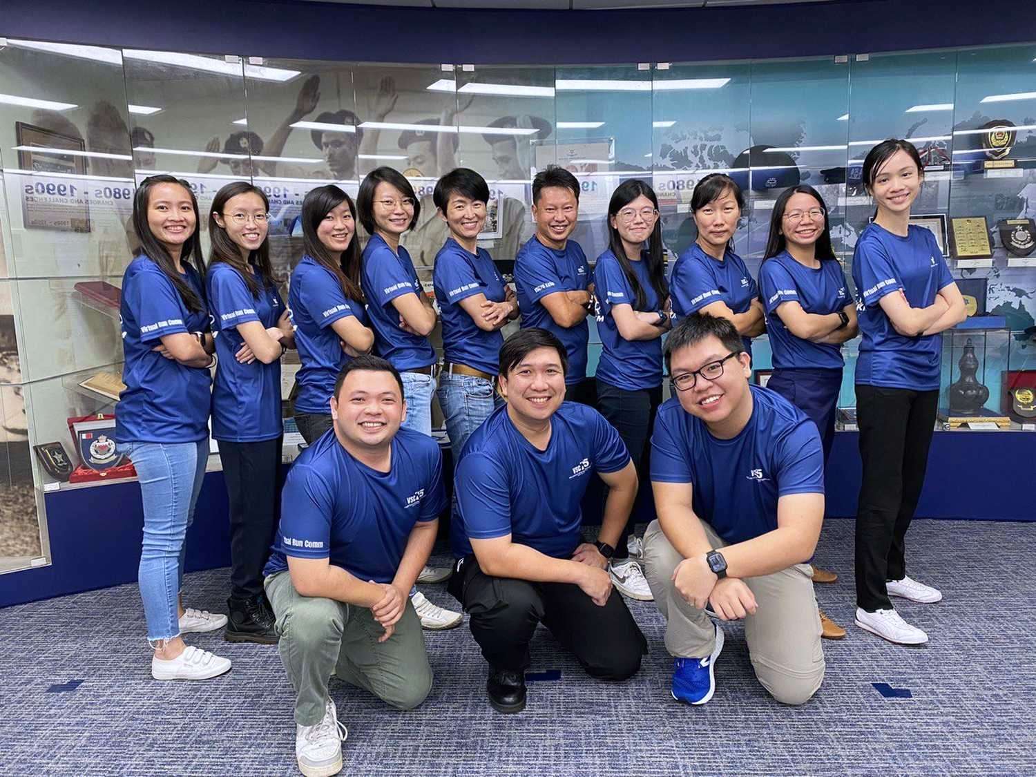 a team of officers wearing blue polo tee posing together with the men kneeing infront and the women at the back row posing with their arms crossed