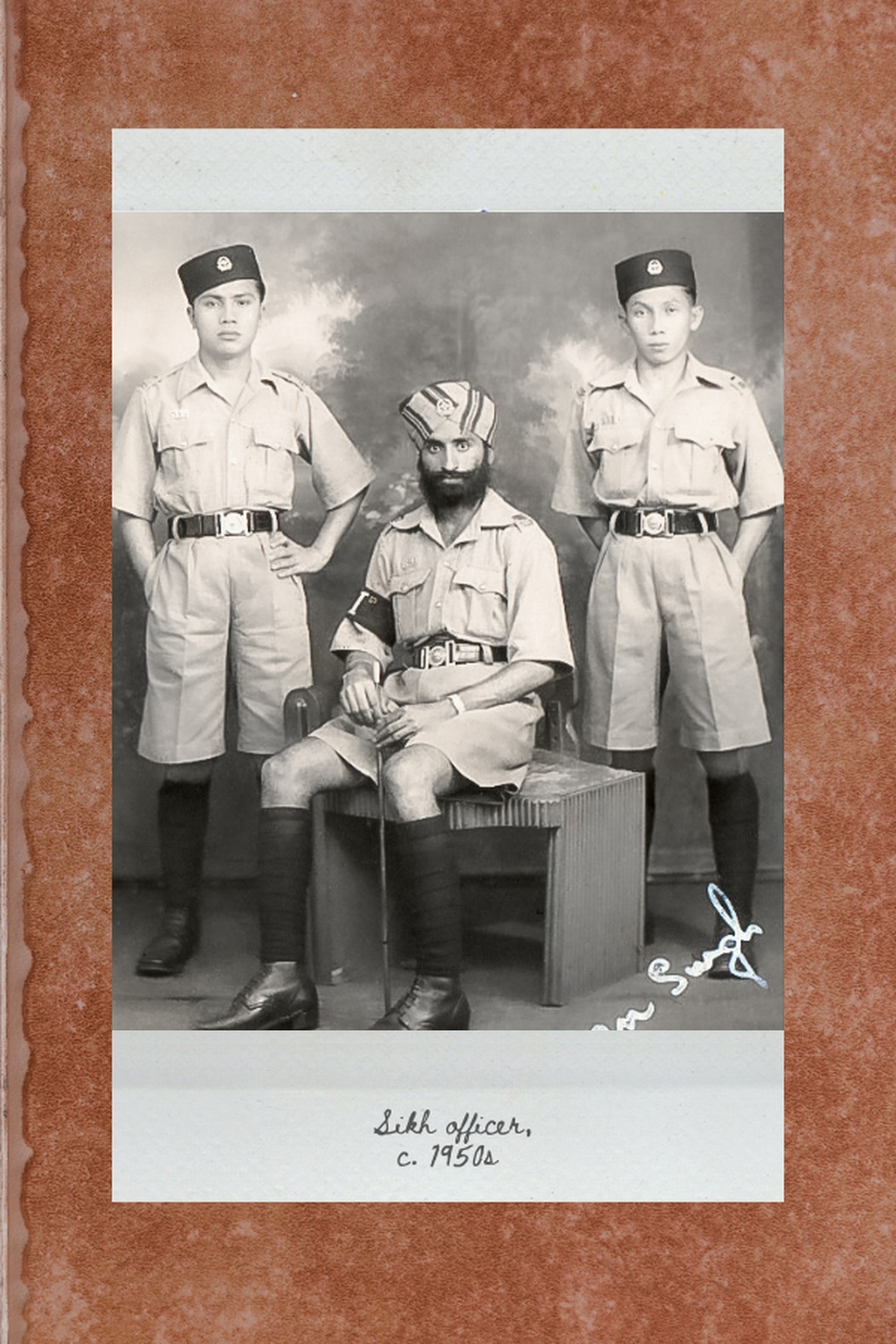 a sikh officer seated in uniform with a training pace stick while two malay officers are standing behind him