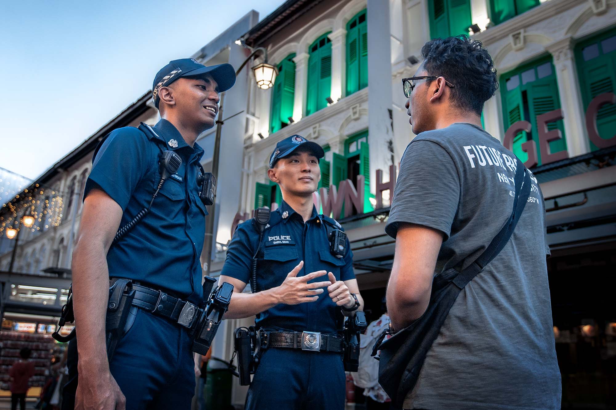 two male police officers interacting with a member of the public, wearing a grey t-shirt and carrying a black sling bag. 