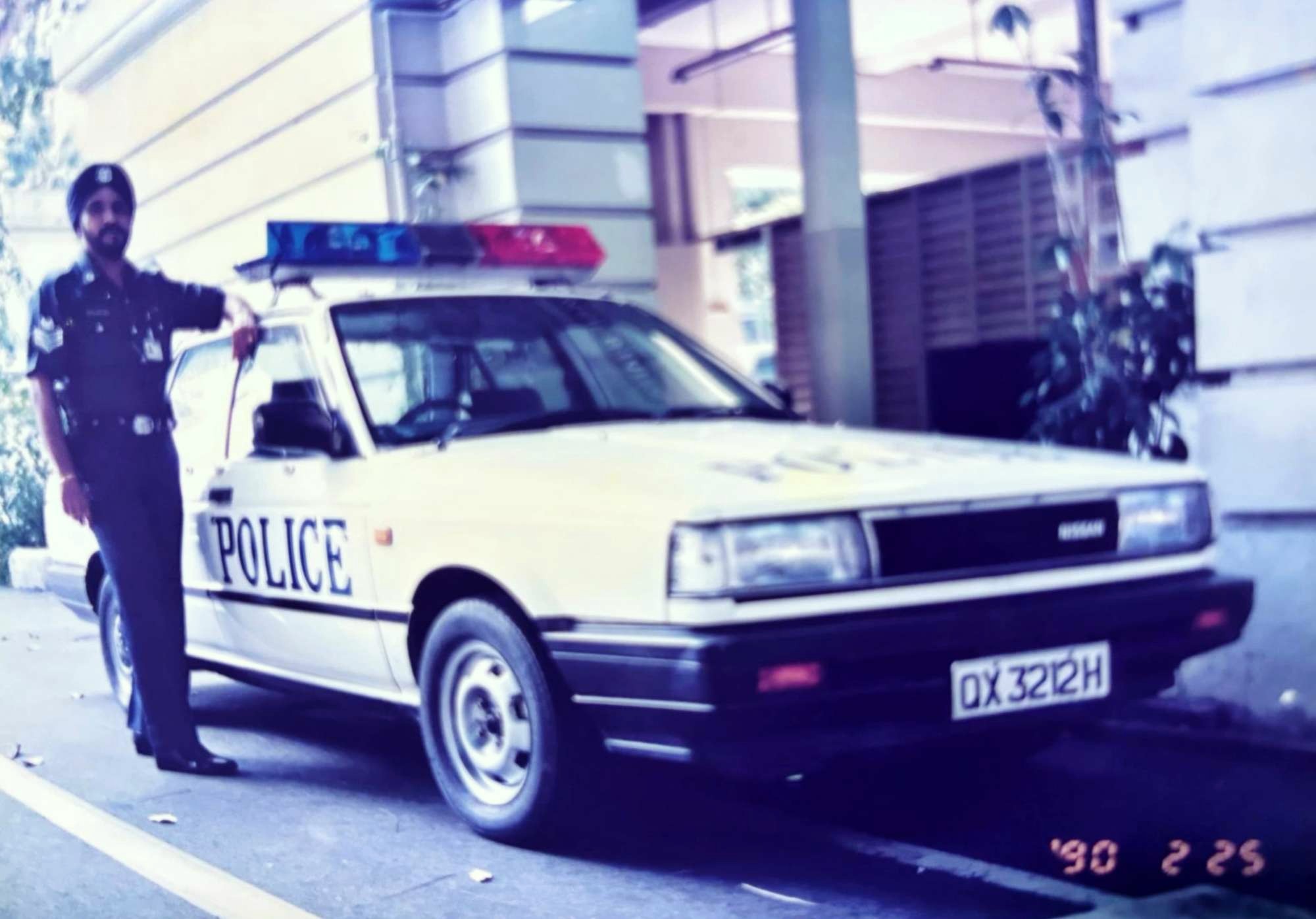 Gurdial in uniform standing infront of the central police station entrance and beside an old police car