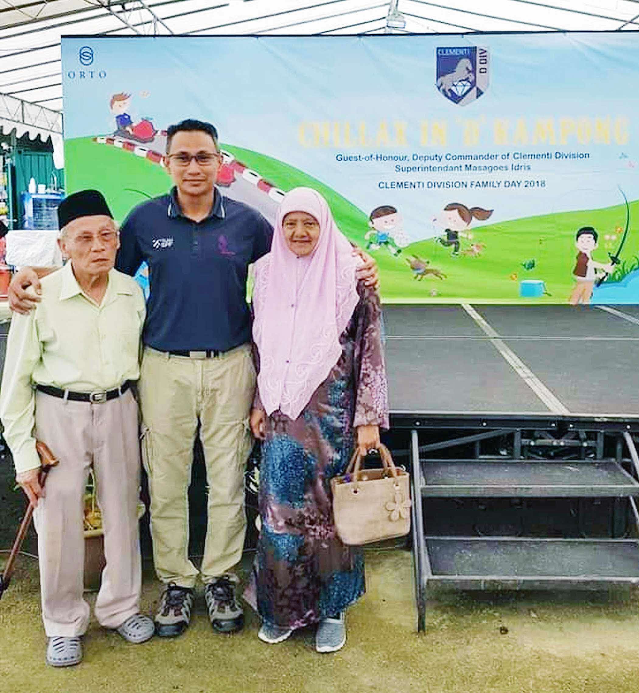 commander with his mum and dad at a community event. his father is wearing a light green shirt and standing on his right while his mum is wearing a pink traditional malay outfit and is on his left.