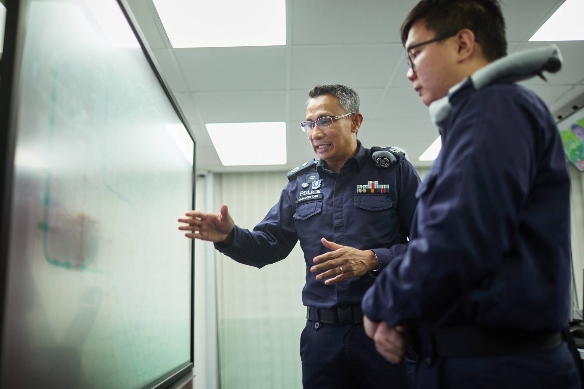 commander masagoes is in the background explaining to an officer on the foreground. He is pointing to a board on the left